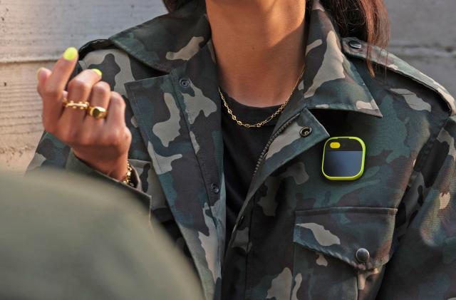 A cropped image showing the torso of a women wearing a camouflage jacket with a yellow Humane AI Pin on the chest.