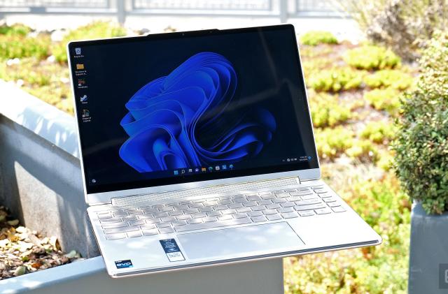 The Lenovo Yoga 9i laptop in white sits open and facing the camera as it sits on the edge of an outdoor planter, with lots of plants and bushes in the background.
