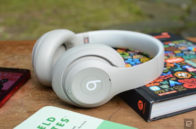 A white pair of Beats Studio Pro over-ear headphones sit on a wooden tabletop as it leans on a hardcover book.