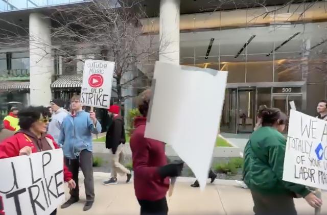 A picket line of strikers protesting outside of Google’s Austin, TX offices. Various people marching outdoors, holding picket signs demanding better treatment.