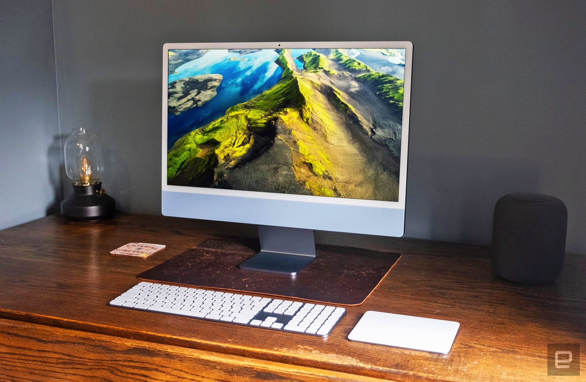 The 2023 Apple iMac with a 24-inch screen and the M3 chip in ice blue sits on a worn wooden corner desk next to a small lamp and an Apple HomePod.